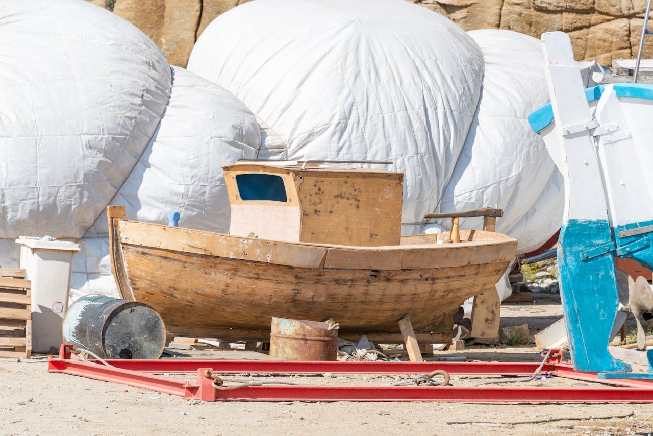 Wooden fishing boat alongside covered boats in a sunny harbor repair area.